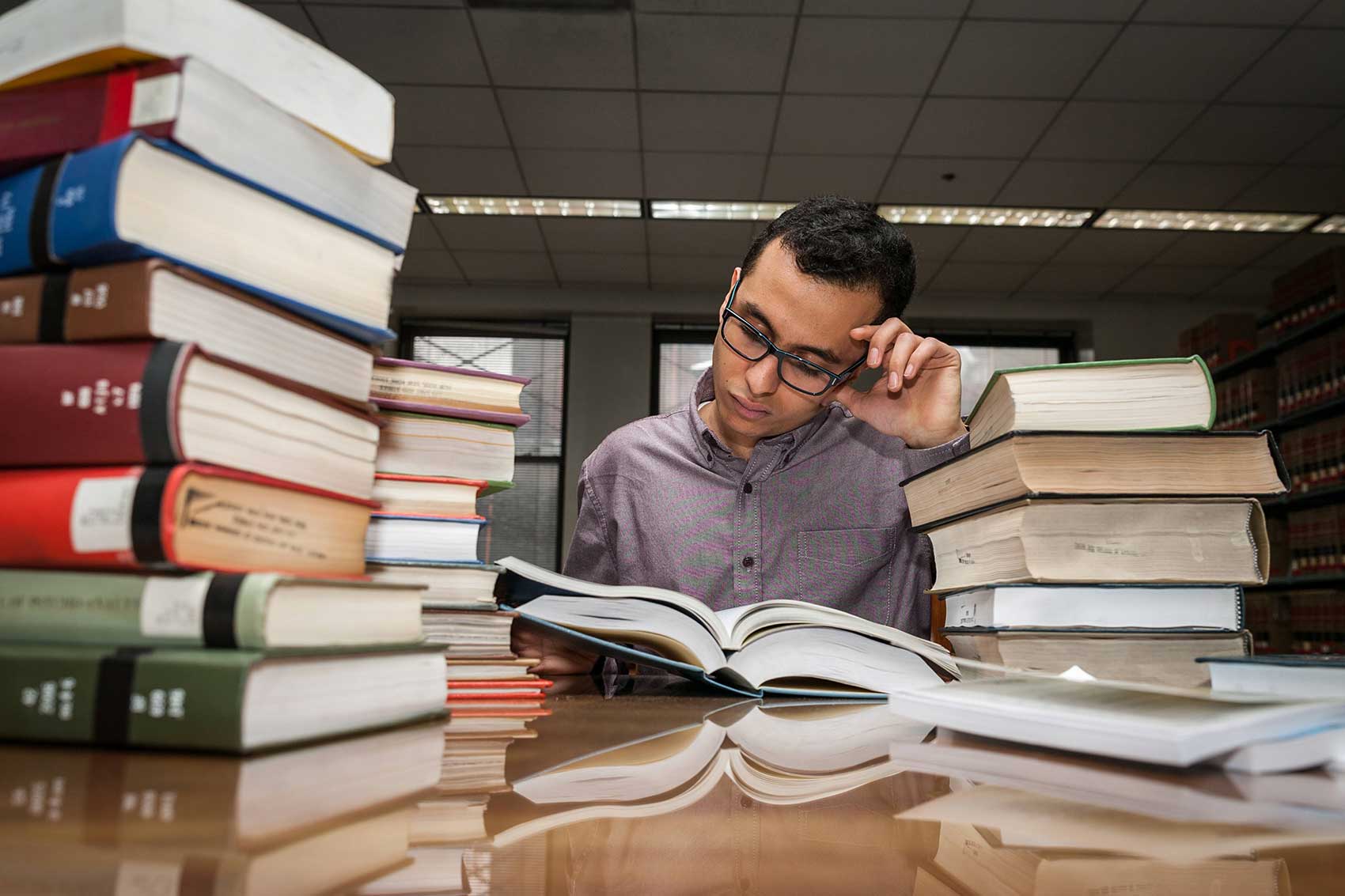 A young man wearing glasses studying in a library, surrounded by large stacks of books.